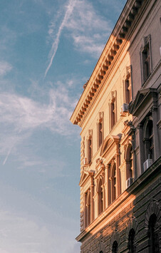 Upward angled view of a single historic building's ornate facade during golden hour, positioned on the right side of the frame against a blue sky. The baroque or neoclassical architecture features elaborate window frames with decorative pediments, detailed cornices with repeating brackets, and warm sandstone-colored walls illuminated by sunset light that makes them glow orange-pink. A single aircraft contrail cuts diagonally across the sky with wispy clouds. The dramatic low angle emphasizes the building's architectural grandeur.