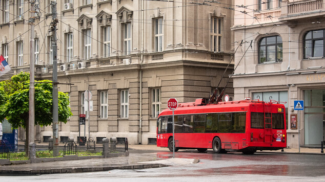 A red electric trolleybus numbered 29 on a wet city street after rain, with overhead power lines visible above. The modern bus has pantograph connectors to the wires. Behind it stand several 19th century European-style buildings in beige and grey stone with classical architectural details. The wet pavement creates reflections. Street infrastructure including traffic signs, a pedestrian crossing sign, and lamp posts with power lines are visible.
