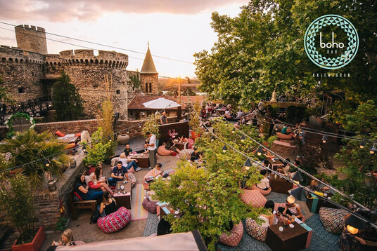 Evening scene at Boho Bar, an outdoor rooftop venue adjacent to medieval fortress walls. Multiple levels of wooden decking hold tables and colorful patterned bean bag seats where numerous patrons socialize with drinks. String lights hang overhead, and lush green trees provide shade. The stone fortress with a conical tower creates a dramatic backdrop. The atmosphere appears relaxed and bustling during golden hour.